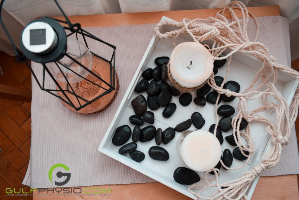 A pair of paraffin wax candles on a tray with black stones on a table right beside an oil lamp