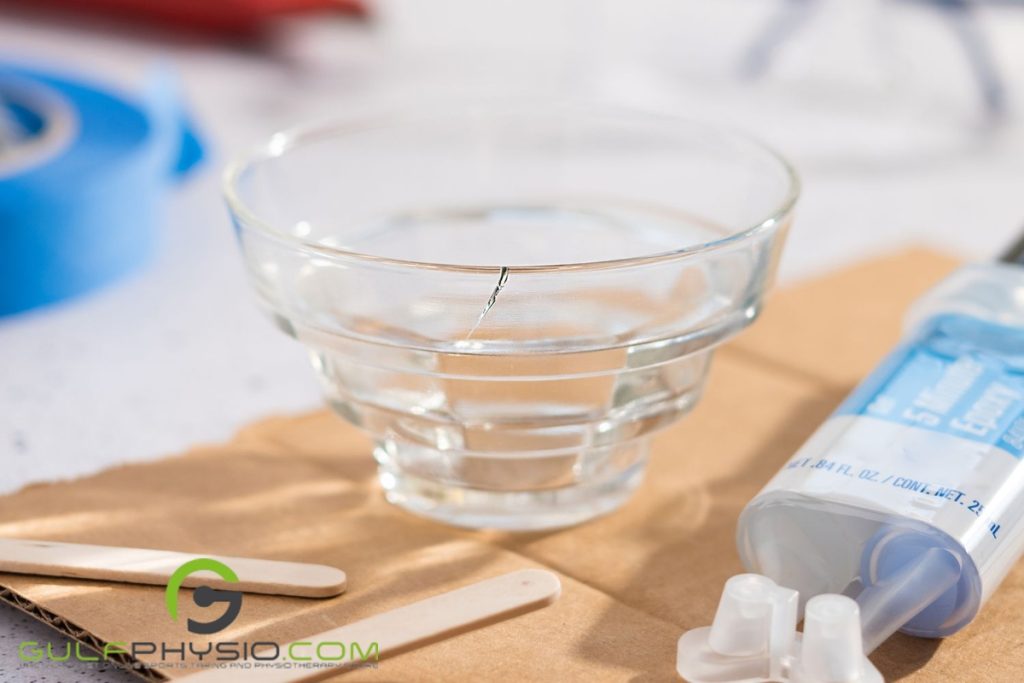 A cracked glass cup on a table surrounded by materials to fix it like paraffin wax