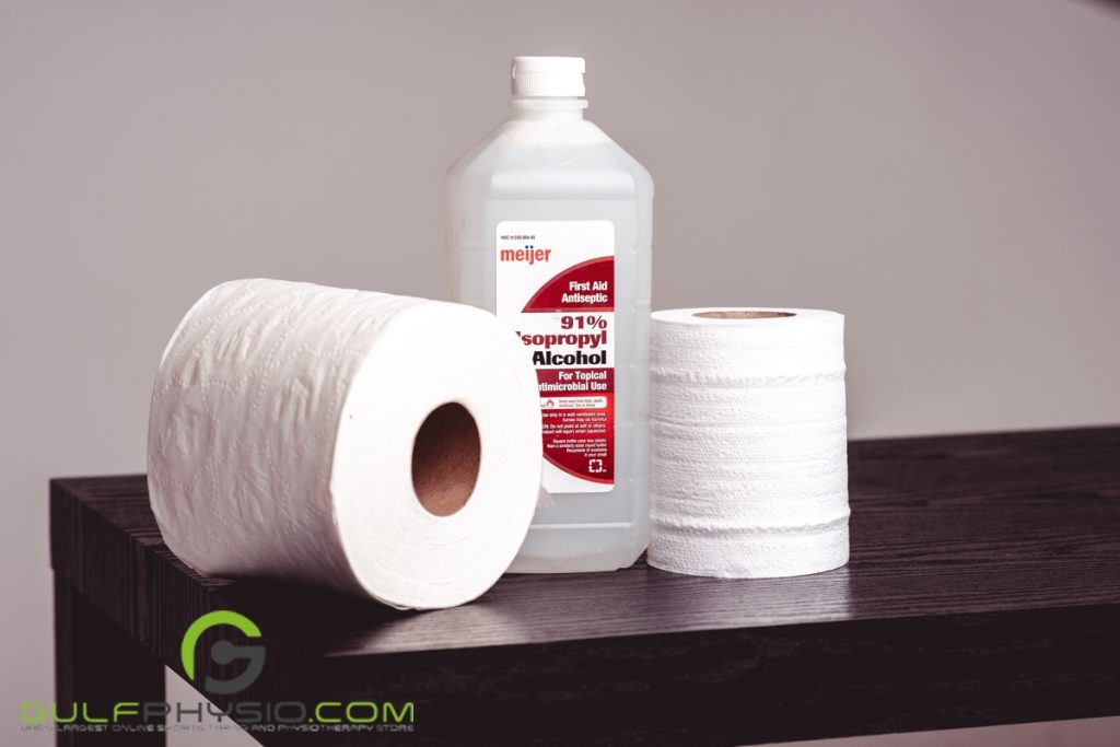 A bottle of isopropyl alcohol is nestled in between two rolls of toilet paper while on top of a wooden table