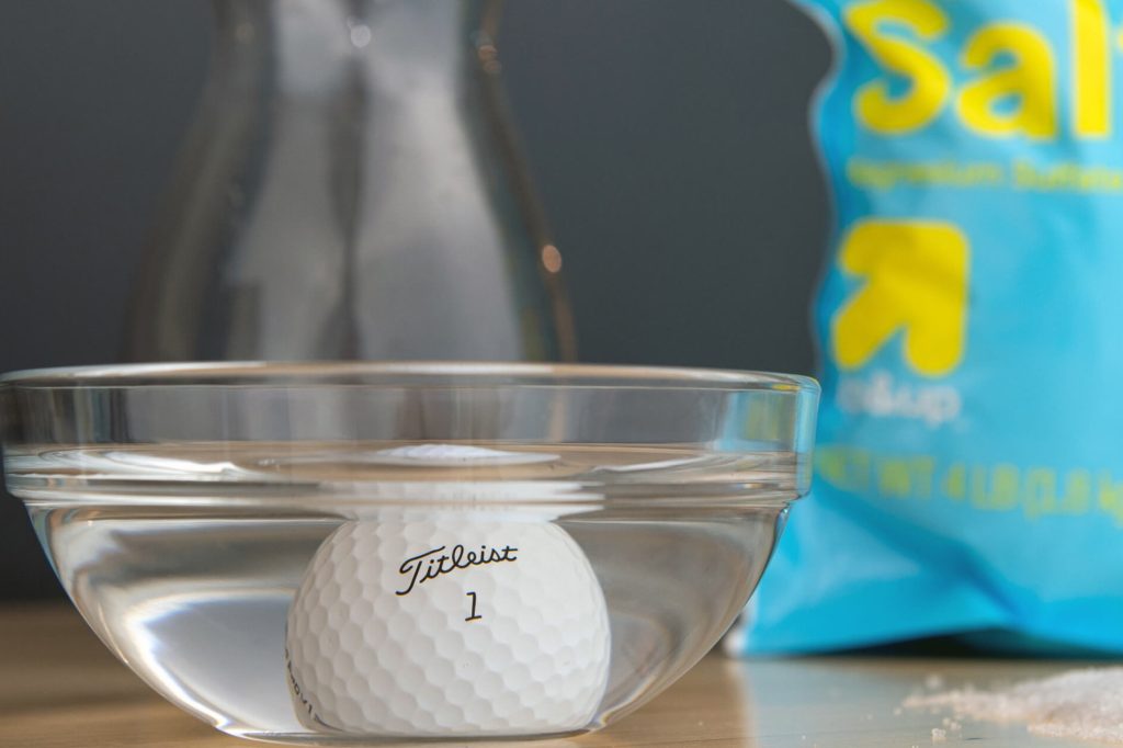 A bowl with a golf ball in a magnesium sulphate solution is on a table surrounded by a canister of water and a bag of Epsom salt