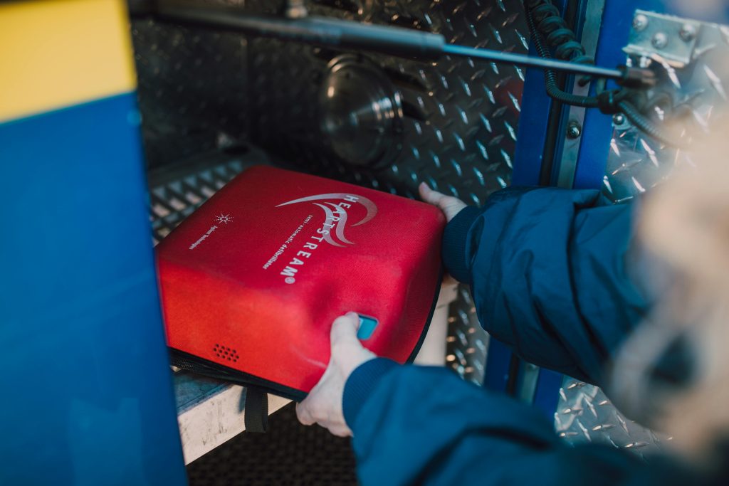 An AED is being taken from the inside of an emergency vehicle by a first responder wearing a dark blue uniform