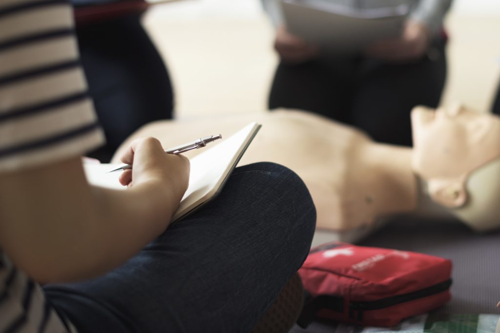 A person is sitting down on the floor while writing down the tips they are learning from an emergency preparedness training session in front of a life support dummy