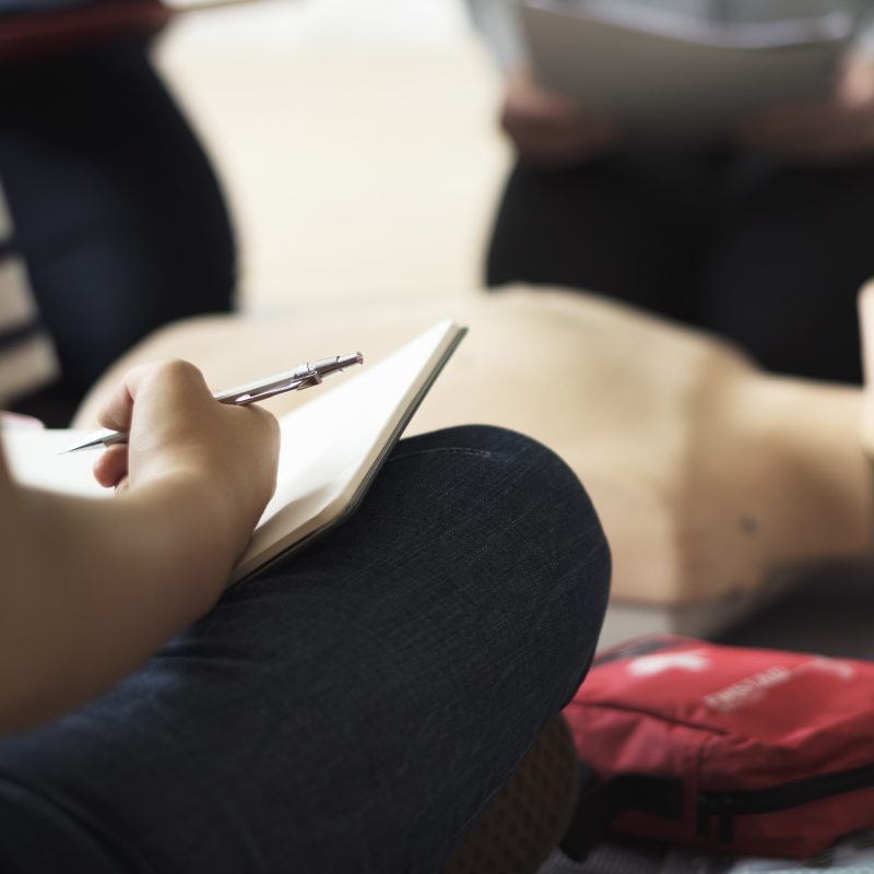 A person is sitting down on the floor while writing down the tips they are learning from an emergency preparedness training session in front of a life support dummy