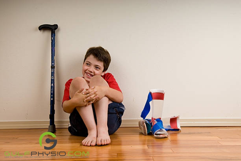 A boy sitting on the floor and hugging his knees while smiling. Beside him is a walking cane and a pair of orthopedic foot braces. 