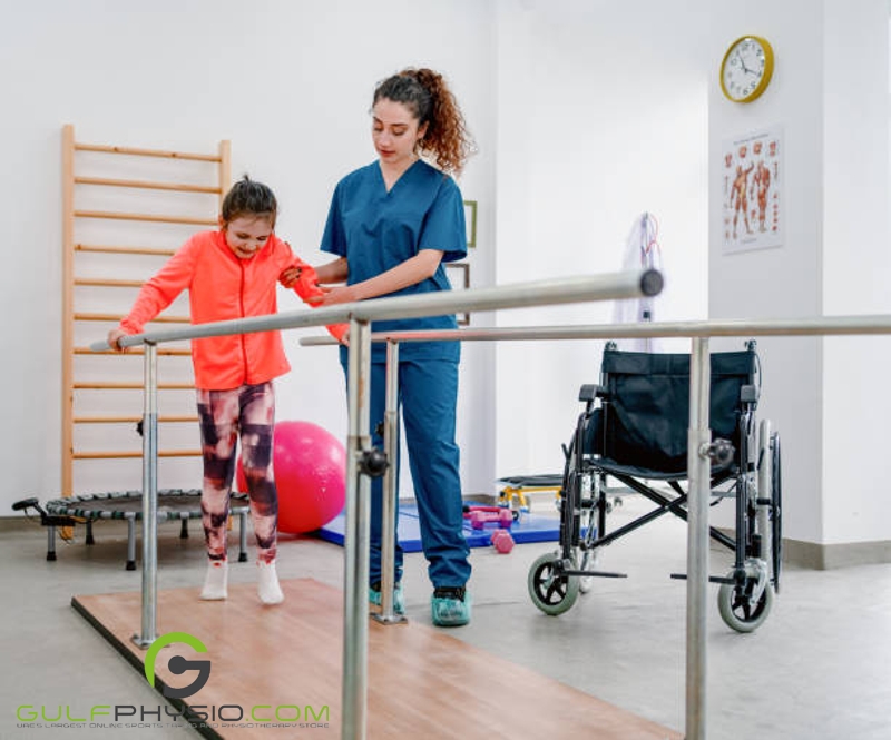 A young girl learning how to walk with the assistance of a physical therapist. They are making use of parallel bars. There is an empty wheelchair in the background.