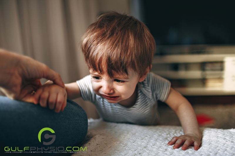 A young boy (around toddler age) struggles to climb up onto a low bed. Off camera an adult hand gently holds his arm.