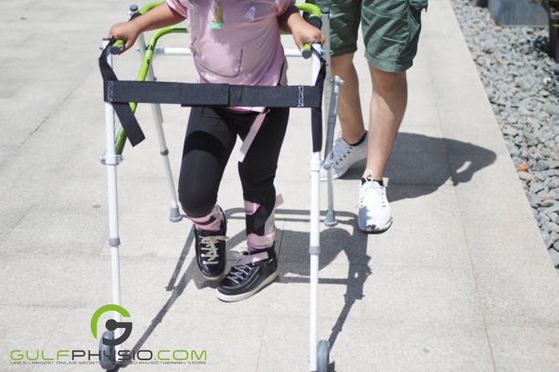 A photo of a young girl using an assistive walker and orthopedic walking braces. An adult is guiding her from behind. 