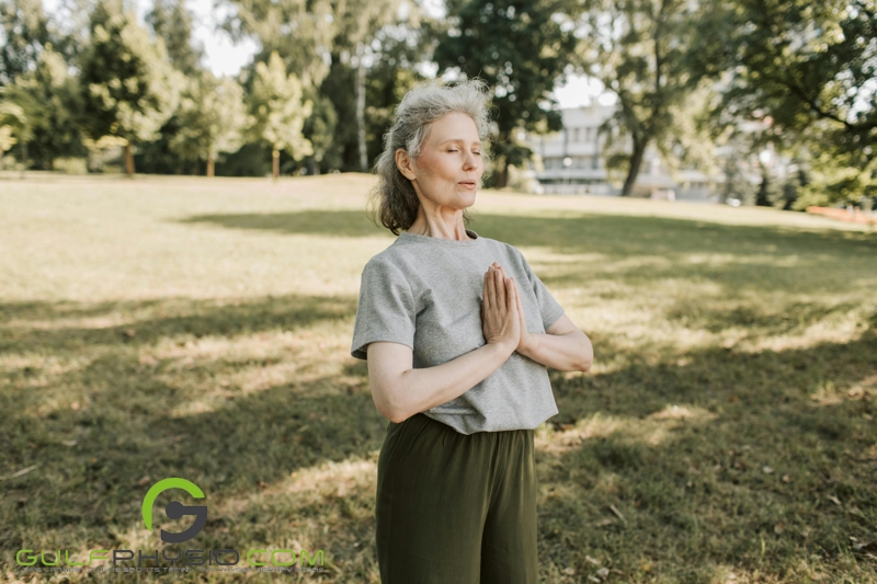 A woman in a park with her hands in front of her chest as she inhales.