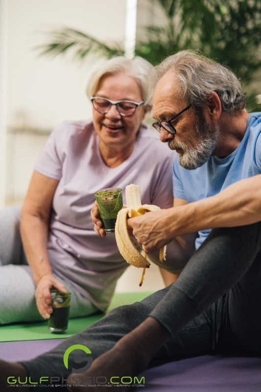 An elderly couple seated on yoga mats, drinking what looks like green smoothies. The man is also eating a banana.