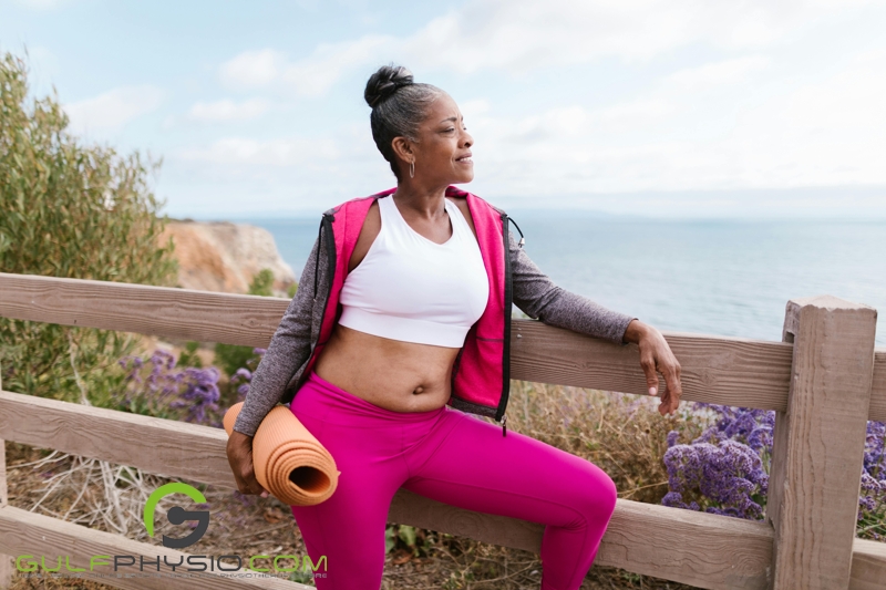 An older woman dressed in athletic wear and holding a yoga matt. She is leaning on a wooden fence and looking out onto the horizon.