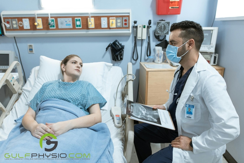 A doctor talking to his patient. The patient is lying down on a hospital bed while the doctor is seated on a stool beside the bed. The doctor is also holding a clipboard containing various medical papers and what appears to be the MRI scan