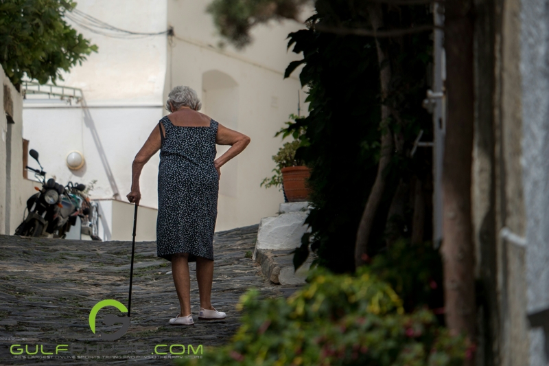  A photo of an older woman walking with a cane. She is walking away from the camera on a slightly uphill path.