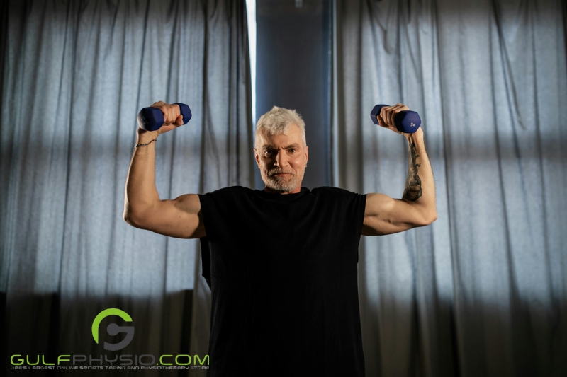 A muscular older gentleman holding small barbells in his hands while he flexes.